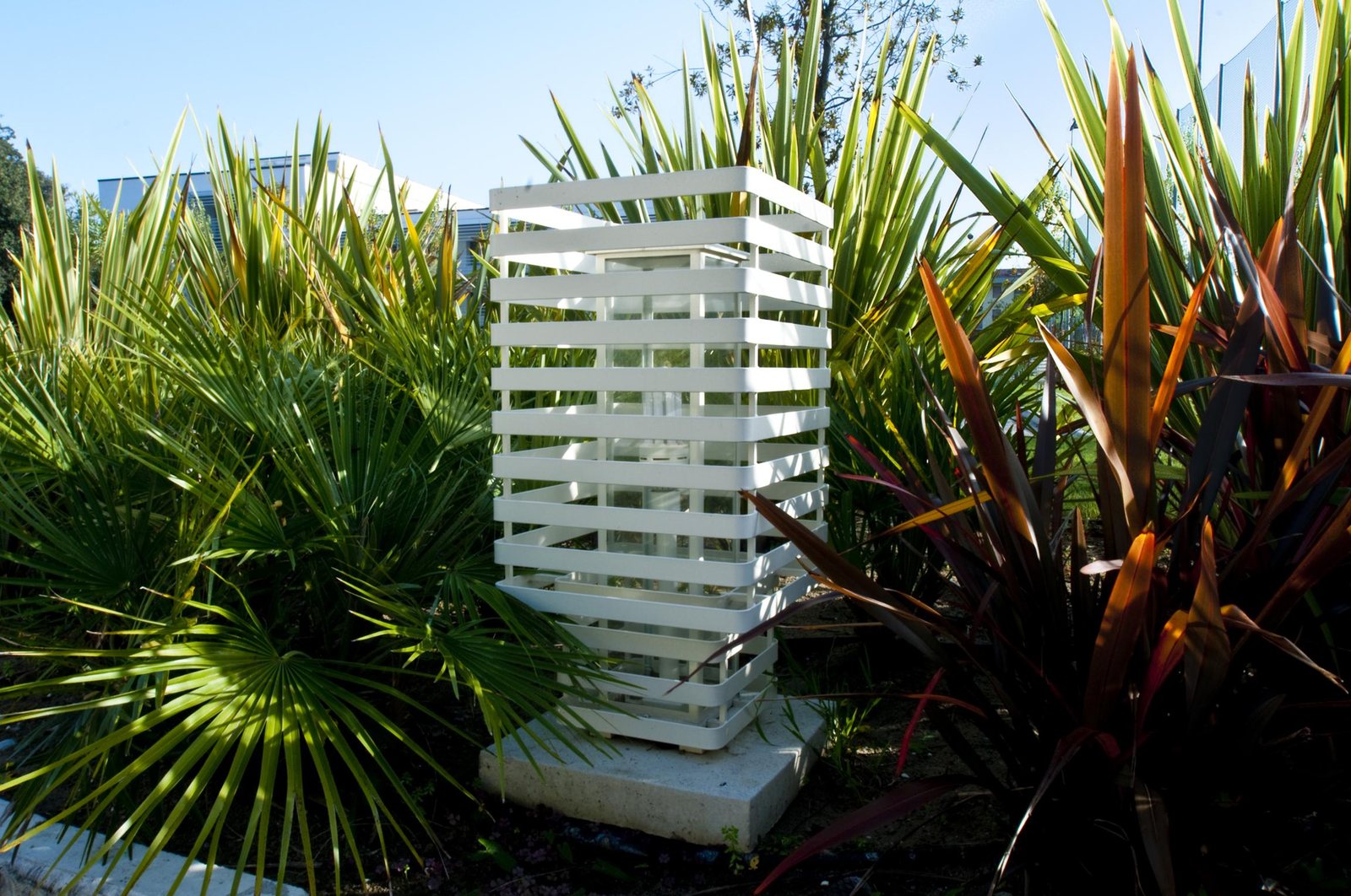 Il paletto a lamelle bianche fra le foglie di palma e le cordyline del giardino