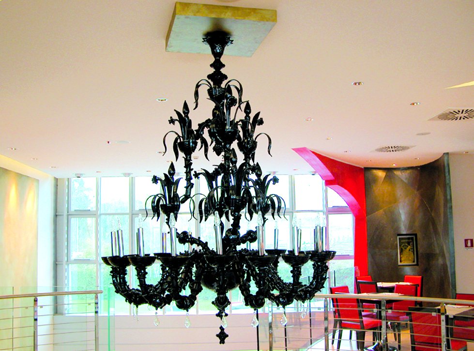 Le lustre noir de style vénitien de l'UNA Hotel Malpensa : bras sur plusieurs rangs et bougies claires, devant la baie vitrée et les chaises rouges