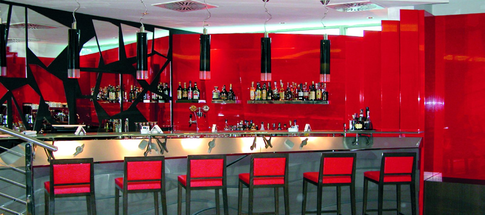 Le bar de l'UNA Hotel Malpensa : une rangée de suspensions cylindriques noires en verre de Murano au-dessus du comptoir en métal, entre le mur rouge et les tabourets rouges