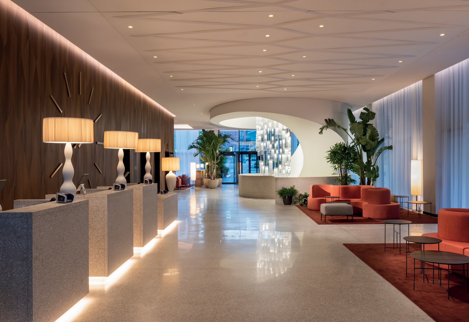 The lobby of the Mövenpick Hotel Basel: table lamps with a sinuous white base and a lit pleated shade on the reception desks, coral sofas on the right