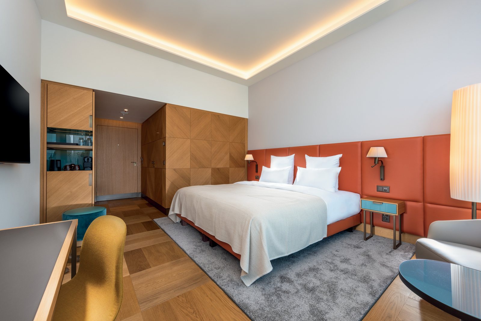 A king room at the Mövenpick Hotel Basel: wall lamps with pleated shades and reading spots on the orange headboard, a lit floor lamp at the right edge