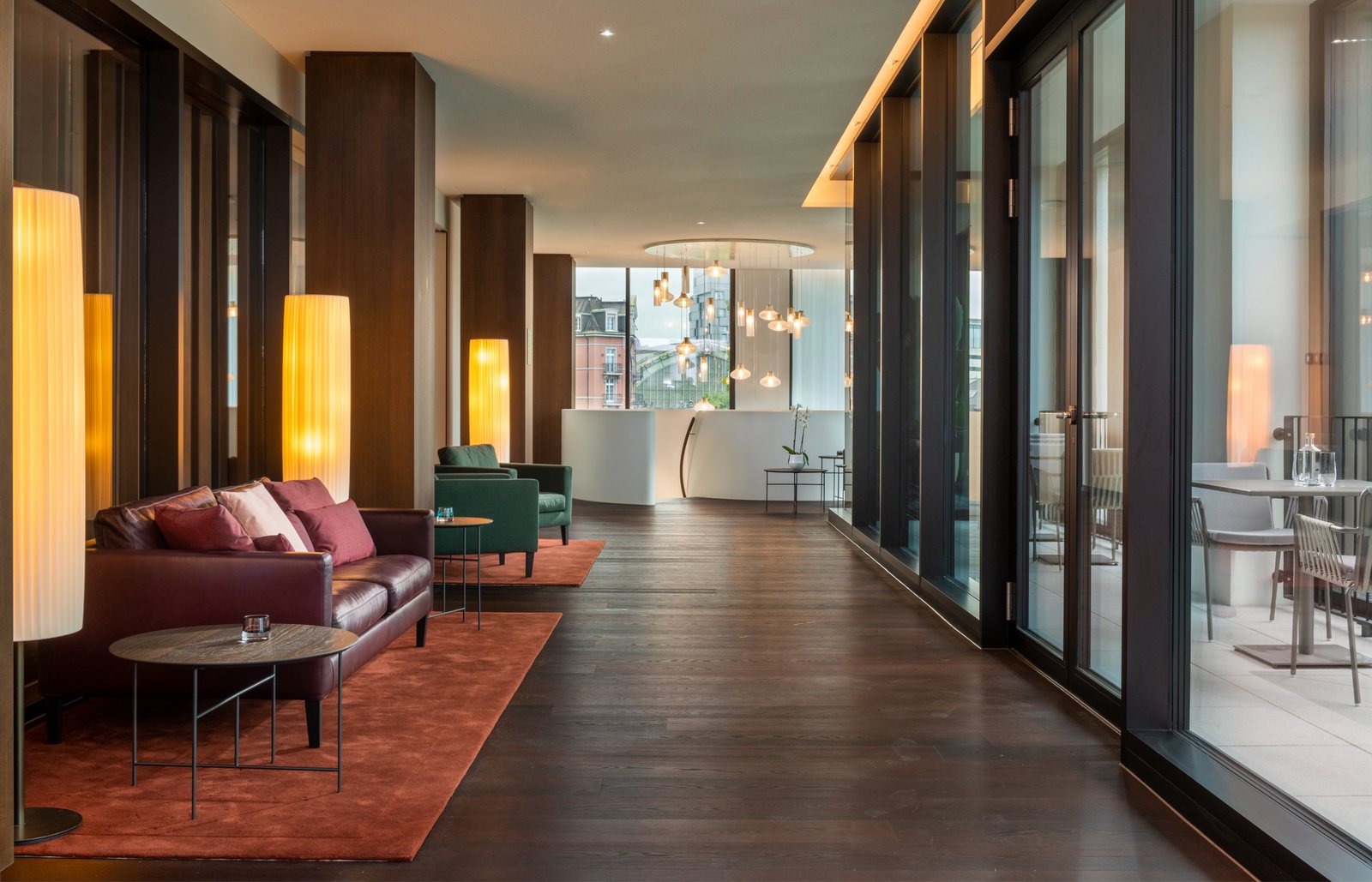 The lounge of the Mövenpick Hotel Basel: a row of lit floor lamps with pleated cylindrical shades beside the dark pillars, a burgundy sofa and glass doors onto the terrace