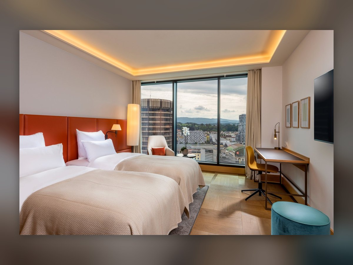 A twin room at the Mövenpick Hotel Basel with full-height glazing over the city: wall lamp on the orange headboard, lit floor lamp in the corner, brass-coloured lamp on the desk