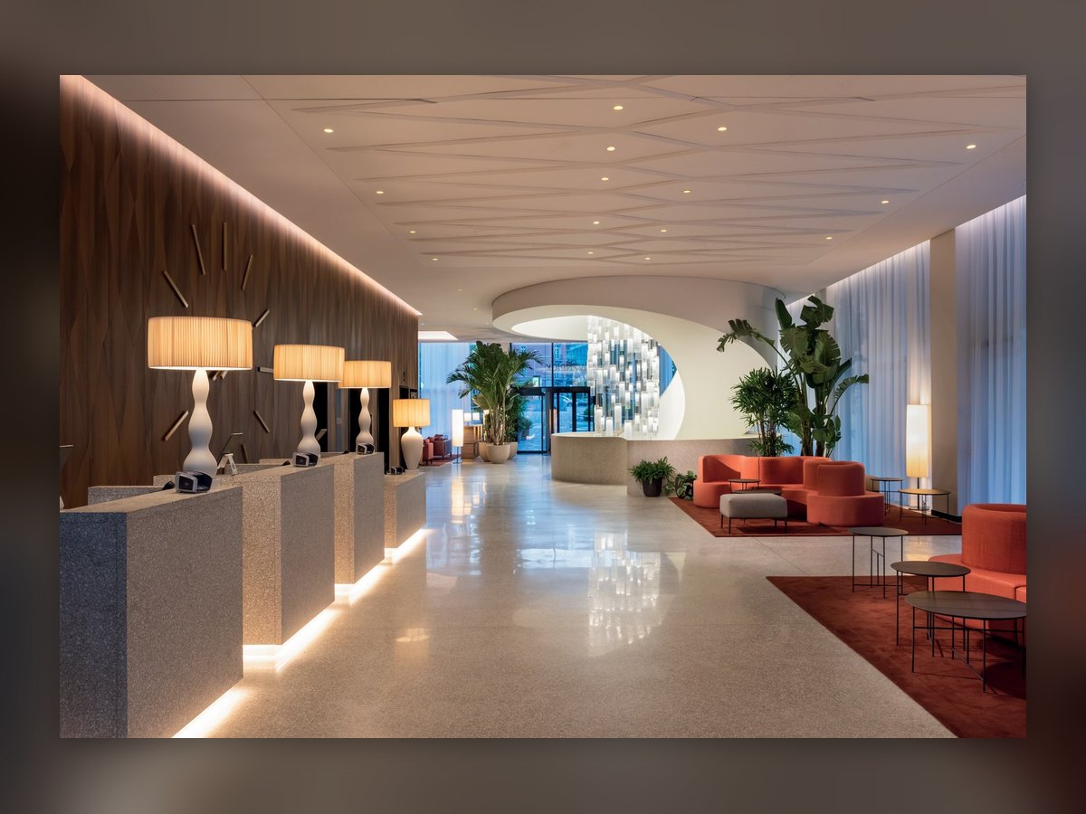 Le hall du Mövenpick Hotel Basel : sur les comptoirs de la réception, des lampes de table au pied blanc sinueux et à l'abat-jour plissé allumé, canapés corail à droite
