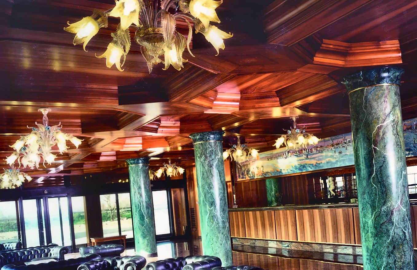La hall del Russott Hotel di Mestre: lampadari veneziani a foglie e fiori color paglierino accesi sotto il soffitto di legno a cassettoni, colonne verdi venate e poltrone di pelle nera