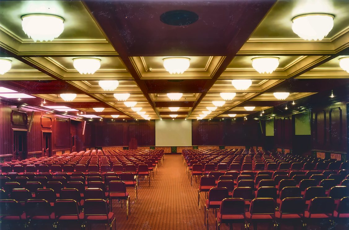 Il salone congressi del Russott Hotel di Mestre: plafoniere circolari in vetro di Murano accese nei cassettoni chiari del soffitto, file di sedie rosse e pareti a pannelli di legno