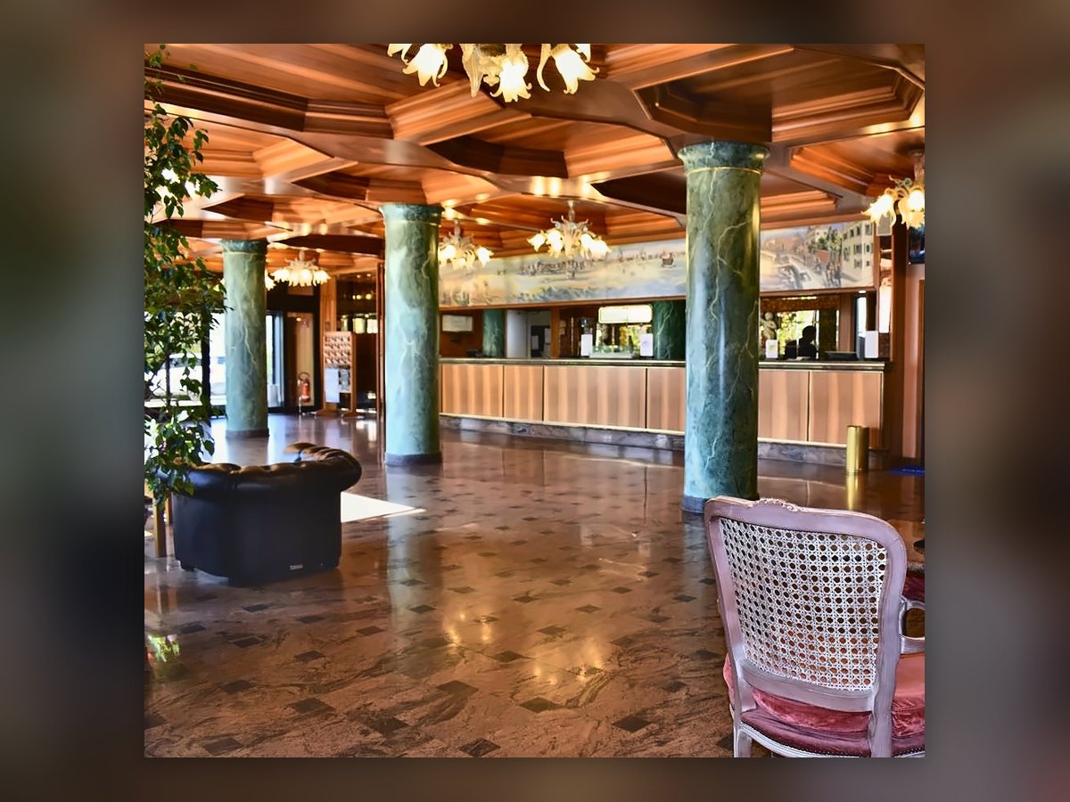 La hall del Russott Hotel di Mestre con i lampadari veneziani accesi sopra le poltroncine di paglia di Vienna rosa e le colonne verdi, il banco della reception sullo sfondo
