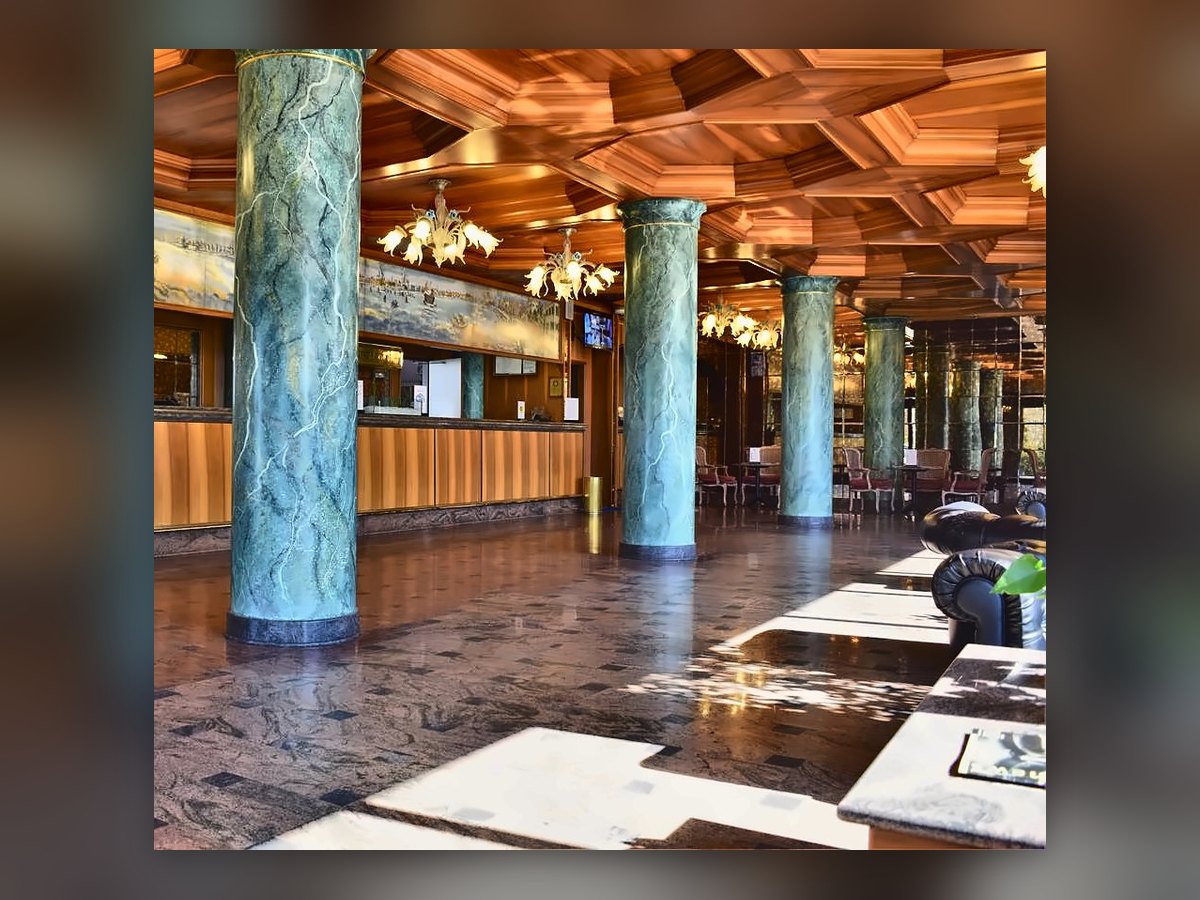 Il banco della reception del Russott Hotel fra le colonne verdi venate, con due lampadari veneziani accesi e la luce del sole sul pavimento di granito