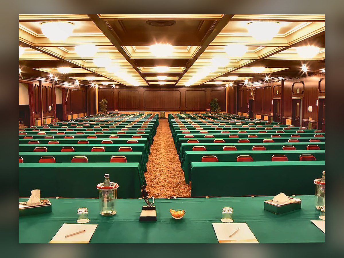 Il salone congressi del Russott Hotel allestito per un convegno: plafoniere circolari accese nei cassettoni, file di tavoli coperti di verde e sedie rosse