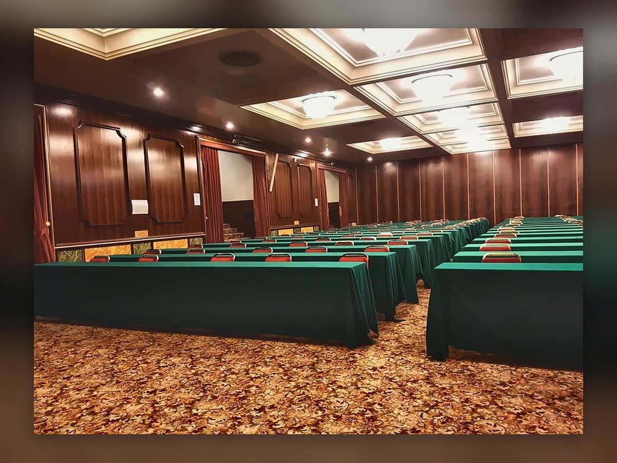 Una sala del centro congressi del Russott Hotel vista di lato: plafoniere circolari accese nei cassettoni del soffitto, tavoli verdi in file e pareti di legno con tende rosse