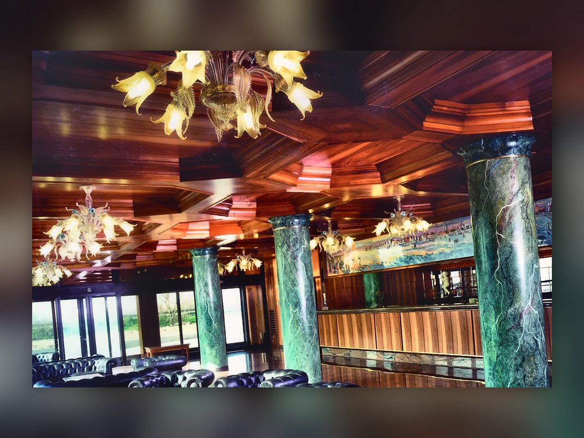 Le hall du Russott Hotel à Mestre : lustres vénitiens couleur paille à feuilles et fleurs allumés sous le plafond à caissons en bois, colonnes vertes veinées et fauteuils en cuir noir